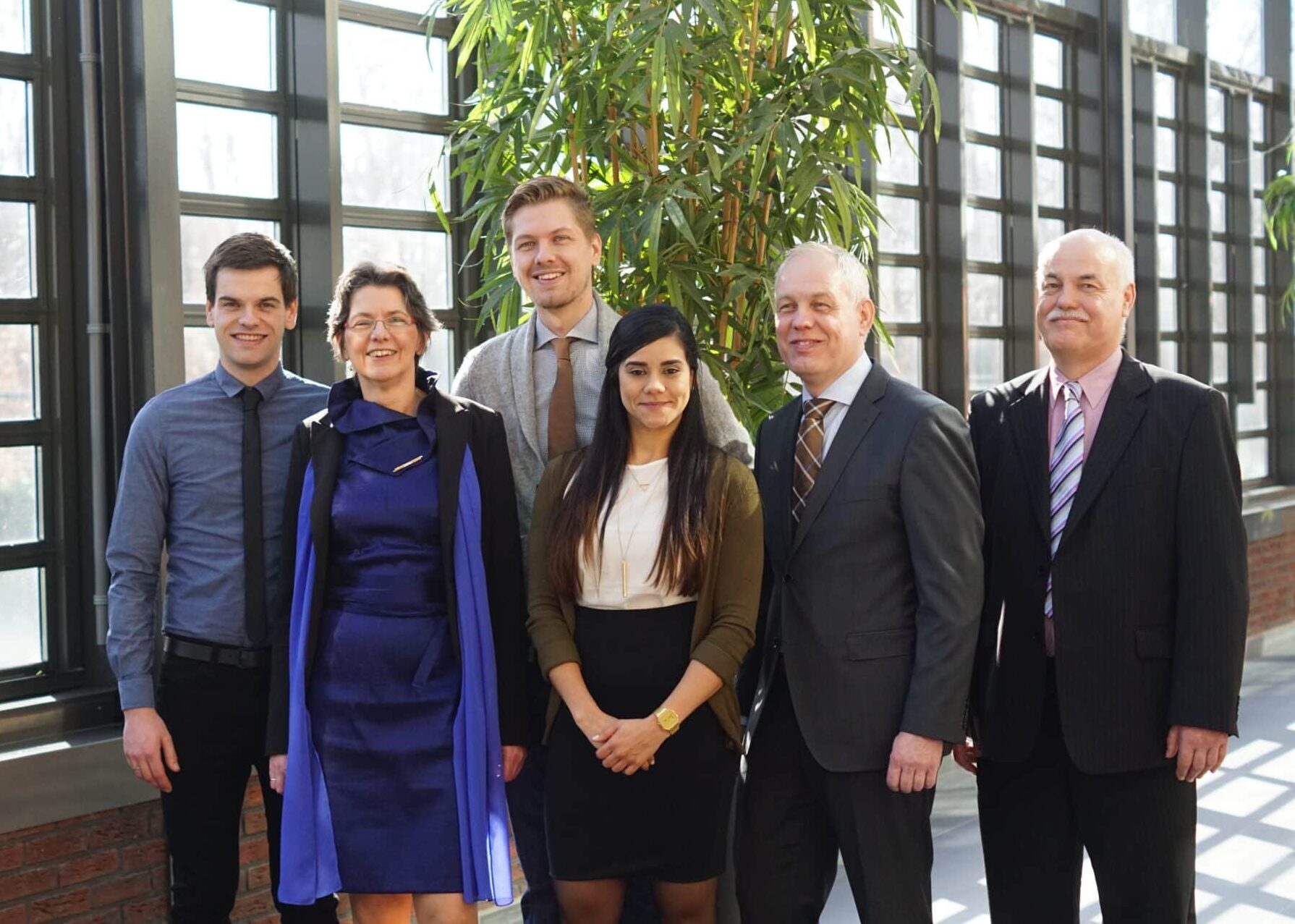 Groep mensen in formele kleding poseert samen binnen voor grote ramen en groen decor.